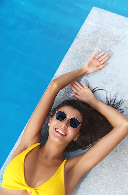 Toerisme en spa bovenaanzicht van sexy brunette vrouw in bikini en zonnebril liggend in de buurt van zwembad vrouwelijk model zonnebaden in het zwembad en glimlachend in de camera