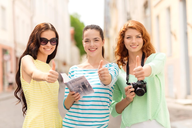 toerisme, reizen, vrije tijd, vakantie en vriendschapsconcept - glimlachende tienermeisjes met stadsgids en camera die duimen in openlucht tonen