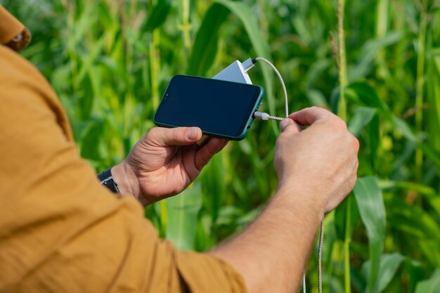 Toerist heeft een smartphone met een draagbare oplader in zijn handen. Man met een powerbank laadt de telefoon op tegen de achtergrond van het maïsveld.