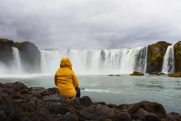 Toerist in een gele jas die ontspant bij de Godafoss-waterval in IJsland