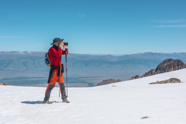 Toerist in rode wandelingen op sneeuwberg nabij afgrondrand op grote hoogte onder blauwe lucht op zonnige dag Man met camera op besneeuwde berg nabij afgrondrand met uitzicht op grote bergketen in de verte