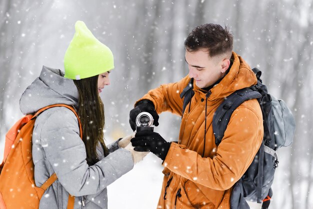 Toeristenjongen met een oranje jasje schenkt hete thee uit thermos in de thermobeker voor meisjes in het koude winterwoud