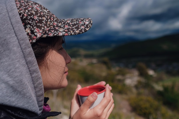 Toeristisch meisje met een pet houdt een mok met een warme drank in haar handen en geniet van het landschap