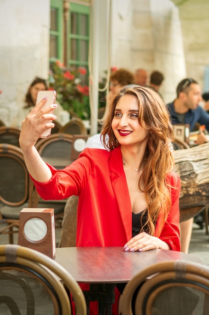 Toeristische jonge blanke vrouw in een rood jasje met koffer neemt een selfie aan de tafel in café buiten.