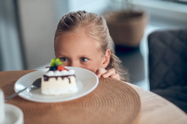 Foto toetje. een schattig blond meisje dat vooruitkijkt terwijl ze naar het plakje cake kijkt