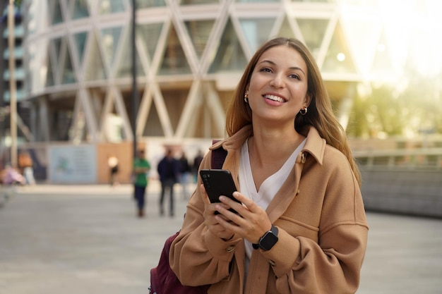 Toevallig vrolijk meisje dat loopt en een smartphone vasthoudt in de straat in de stad en een jas draagt in het voorjaar Stadslevensstijlmensentechnologie