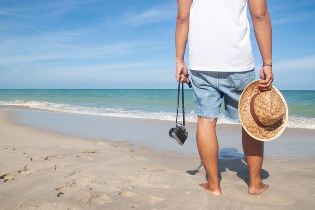 Toevallige stijlmens met camera en hoed die op het strand, Gelukkige vakantie lopen