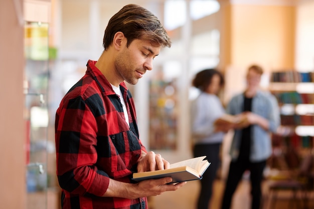 Toevallige student die met boek taken of tekst bekijkt vóór les of examen in universiteitsbibliotheek