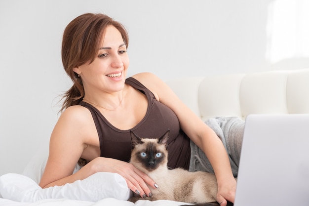 Toevallige vrouw die laptop met haar Siamese kat in witte slaapkamer met behulp van