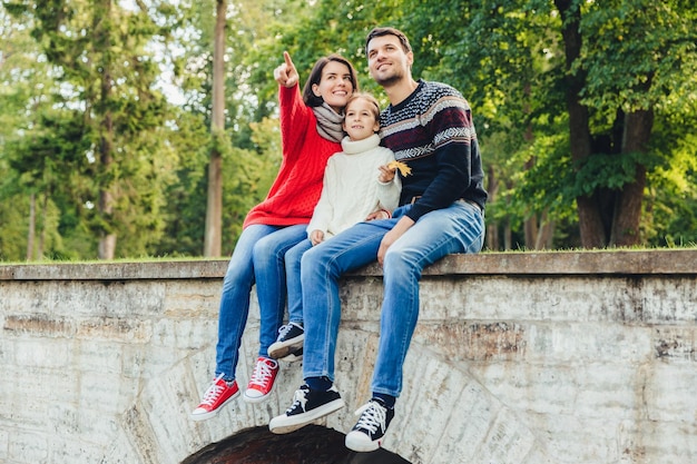 Togetherness and family concept Friendly father mother and daughter sit on stone bridge look from the top notice beautiful rainbow on sky indicate on it with fingers enjoy nature landscapes