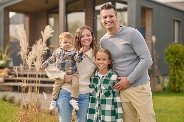 Insieme giovane uomo adulto donna con bambino e ragazza con lunghi capelli biondi sorridente guardando la fotocamera in piedi vicino a casa di campagna in una bella giornata