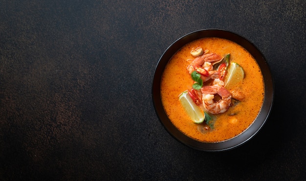 Tom yum kung spicy thai soup with shrimp in a black bowl on a dark stone background top view copy space