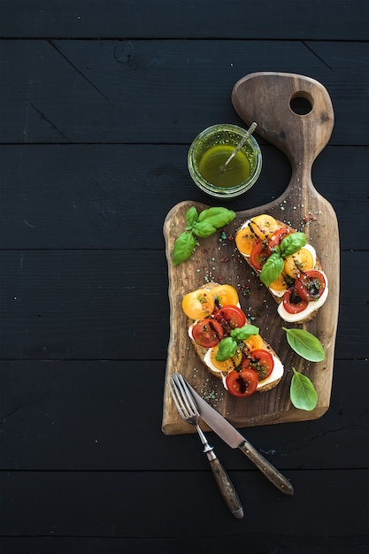 Tomaat, mozzarella en basilicum broodjes op donkere houten snijplank, pesto pot, servies