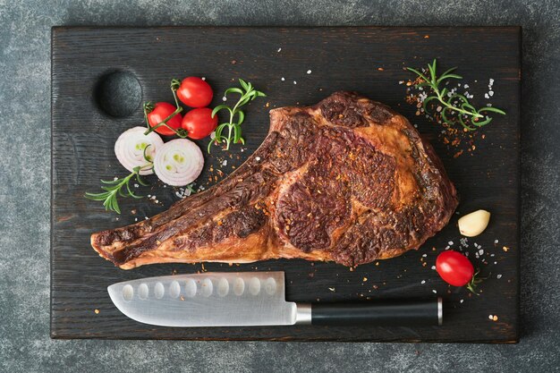 Tomahawk steak Sliced grilled tomahawk beef steak with baked cherry tomatoes herbs and salt on old wooden background Preparing to grill Top view and copy space