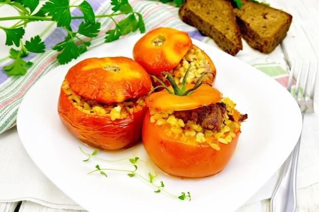 Tomaten gevuld met vlees en gestoomde tarwebulgur in een bord servet vork brood en peterselie op de achtergrond lichte houten planken