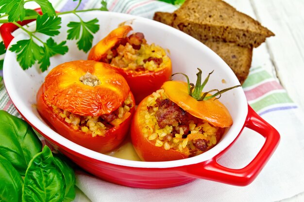 Tomaten gevuld met vlees en gestoomde tarwebulgur in een braadpan op een servet, brood, peterselie en basilicum op een houten plankenachtergrond