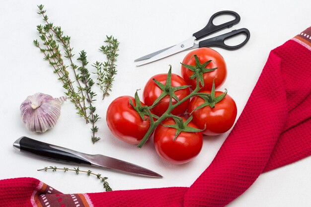 Tomaten op groene tak. Mes en schaar. Takjes knoflook en tijm. Rood servet. Witte achtergrond. Plat leggen