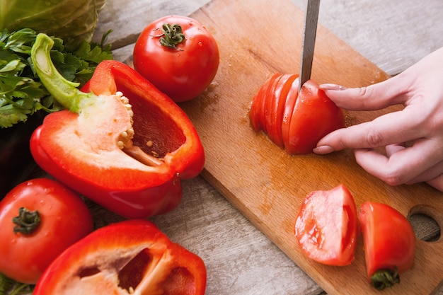 Tomaten snijden op houten bureau door onherkenbare vrouw. Rode peper en peterselie in de buurt op tafel, kookproces van verse groentensalade