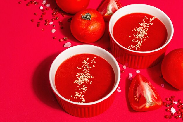 Tomatensoep met sesamzaadjes in kommen op een rode achtergrond. Rijpe groenten, rozen Himalayazout, roze peperkorrels. Een modern hard licht, donkere schaduw, kopieerruimte