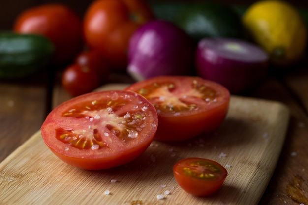 The tomato cut in half is sprinkled with large pieces of salt on a cutting board from a variety of colorful vegetables