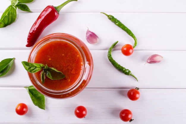 Salsa di salsa ketchup con i pomodori ciliegia e peperoncini, aglio ed erbe roventi in un barattolo di vetro sulla tavola bianca. salsa di pomodoro fatta in casa e pomodori freschi. . vista piana, vista dall'alto