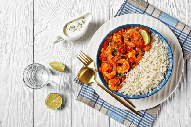 Tomato prawn curry with coriander sprouts and red chili on top served on a plate with jasmine rice and yogurt gravy on a white wooden background with a glass of water and cutlery, top view, close-up