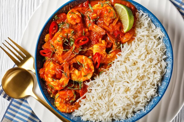 Tomato prawn curry with coriander sprouts and red chili on top served on a plate with jasmine rice and yogurt gravy on a white wooden background with a glass of water and cutlery, top view, close-up