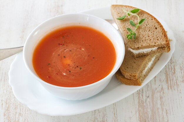 Tomato soup in white bowl with sandwich on white surface