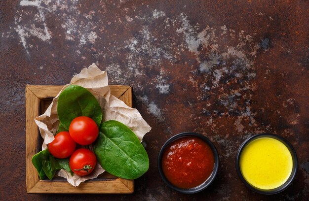 Photo tomatoes and basil leaves in wooden frame with red and yellow sauces on old rusty background, top view, copy space