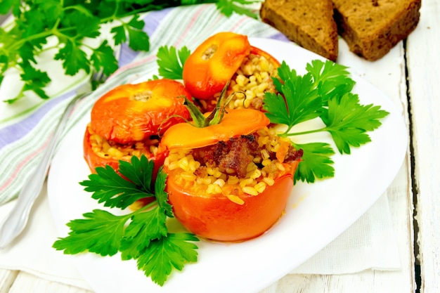 Tomatoes stuffed with meat and steamed wheat bulgur in a white plate, napkin, fork, bread and parsley on the background light wooden boards