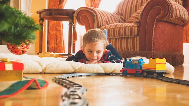 Toned image of smiling little boy playing on wooden floor with his new toy train and railroad. Child receiving presents and toys on New Year or Xmas