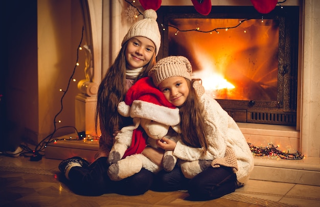 クリスマスイブに暖炉のそばでおもちゃを持って座っている 2 人の幸せな女の子のトーンの古いスタイルの写真