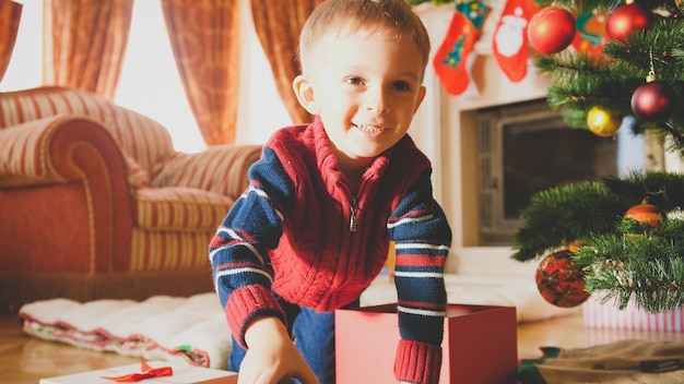 リビングルームの美しい装飾されたクリスマスツリーの下で床を這う幸せな笑顔の小さな男の子のトーンの写真