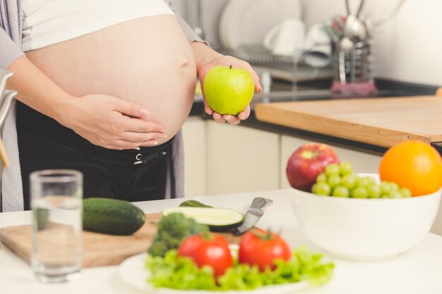 toned photo of pregnant woman posing with green apple while cooking