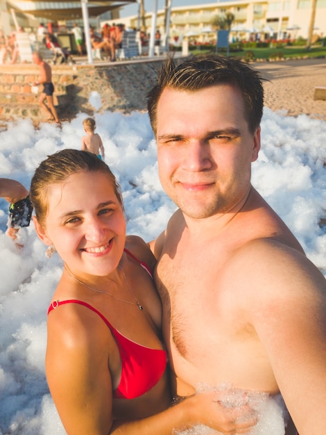 Toned portrait of happy young couple making selfie portrait of the soap foam disco party at sea beach