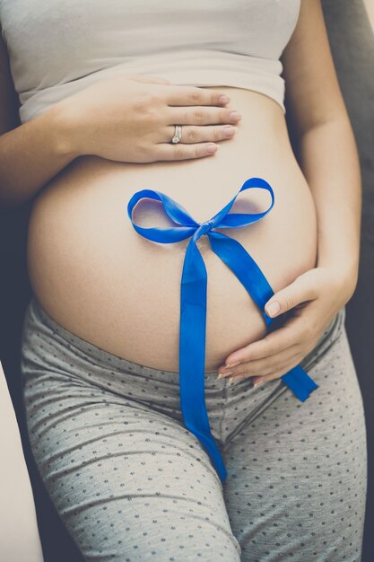 Toned pregnant woman lying on sofa and holding blue ribbon on belly