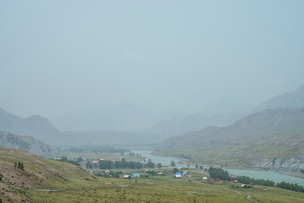 Toneel alpien landschap met bergdorp door bergrivier in mist. Sfeervol mistig landschap met landschap aan de rivieroever. Prachtig uitzicht op het bergdorp aan de rivier bij regenachtig weer.