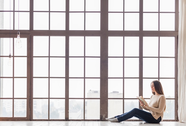 Foto donna delle tenaglie che si siede sul pavimento vicino al libro di lettura della finestra