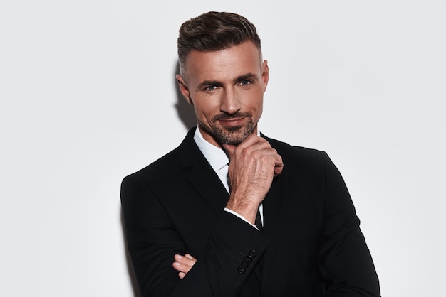 Too good to be real. Handsome young man in full suit keeping hand on chin and looking at camera with smile while standing against white background