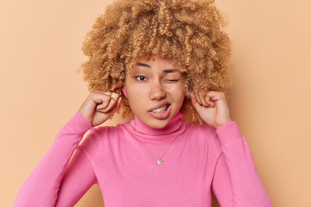 Photo too loud. displeased curly haired young european woman doesnt want to hear something plugs ears ignores noise wears pink jumper isolated over brown background refuses to listen. turn sound off