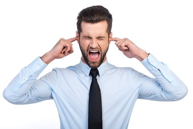 Too loud sound! Frustrated young man in shirt and tie covering ears with fingers and keeping eyes closed while standing against white background