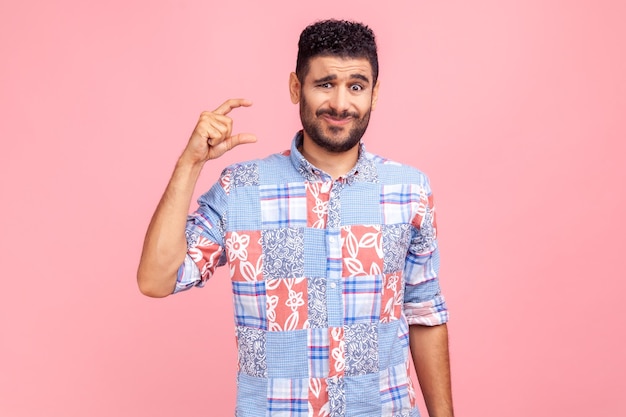 Too small amount. Portrait of disappointed man in blue casual style shirt showing a little bit gesture, dissatisfied with low rating, measuring scale. Indoor studio shot isolated on pink background.