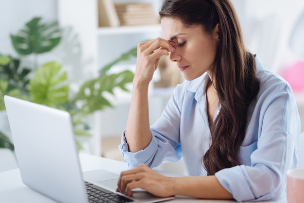 Too tired. Nice pleasant tired woman sitting in front of the laptop and holding her bridge of nose while working late