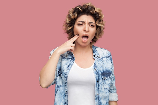 Tooth ache. portrait of sick young woman with curly hairstyle\
in casual blue shirt standing touching her cheek because feeling\
pain on her teeth. indoor studio shot, isolated on pink\
background.