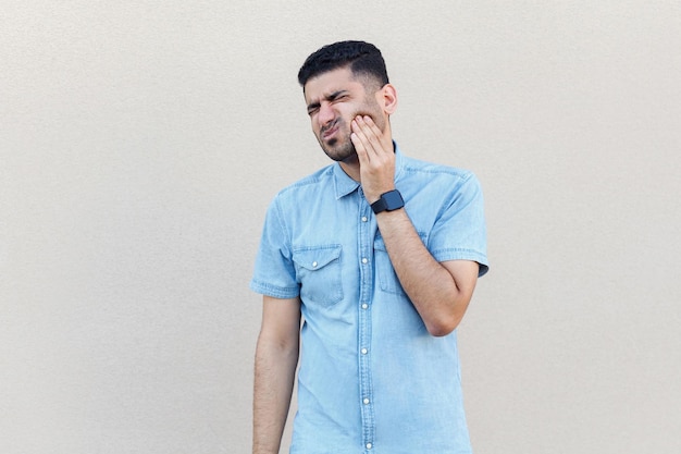 Tooth pain. Portrait of worry sad handsome young bearded man in blue shirt standing, touching his cheek because feeling toothache. indoor studio shot isolated on light beige wall background.