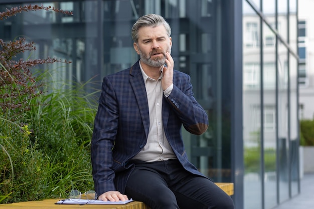 Toothache mature adult male businessman sitting outside office building having severe pain holding