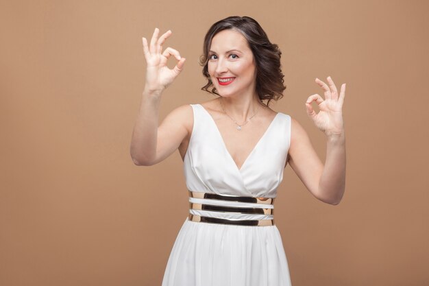 Toothy smiled woman in white dress showing ok sign. Emotional expressing woman in white dress, red lips and dark curly hairstyle. Studio shot, indoor, isolated on beige or light brown background