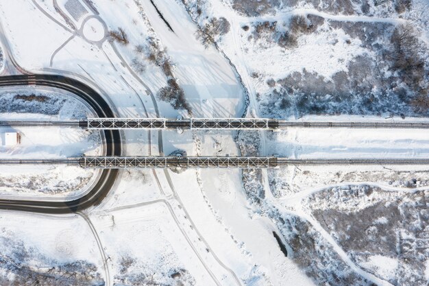 Top aerial view of the railway bridge over the frozen river covered with snow on a winter sunny day.