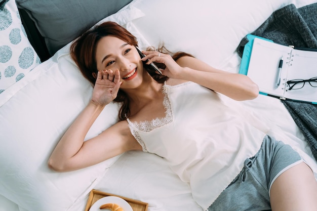 top angle shot cheerful asian girl lying in the bedroom is touching her face with a smile while chatting on the phone in a carefree morning at home.