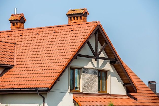 Top of big modern expensive residential house cottage with steep shingled brown roof, high brick chimneys, stucco walls, gutter system and plastic attic windows on blue sky